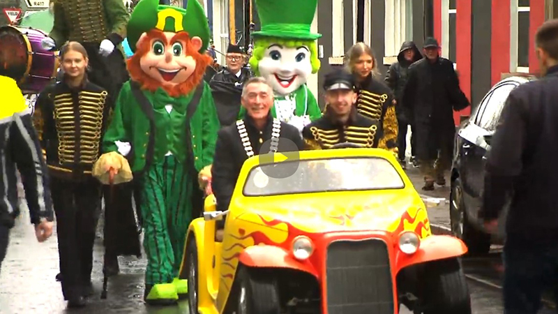 People in festive costumes and band uniforms parade down a street, with a man driving a small colorful car in front